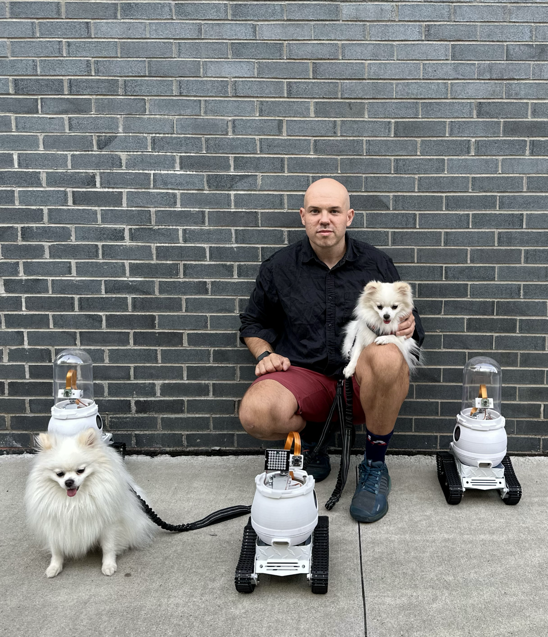 WIM-Z founder kneeling against a brick wall holding a Pomeranian, with prototype rovers beside two dogs